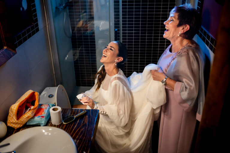 Fotografía de bodas documental: qué es | Adolf Boluda 4 Madre sujetando el vestido de la novia para ayudarla a ir al baño mientras las dos se ríen, boda documental en Villa Mediterráneo, Gandía