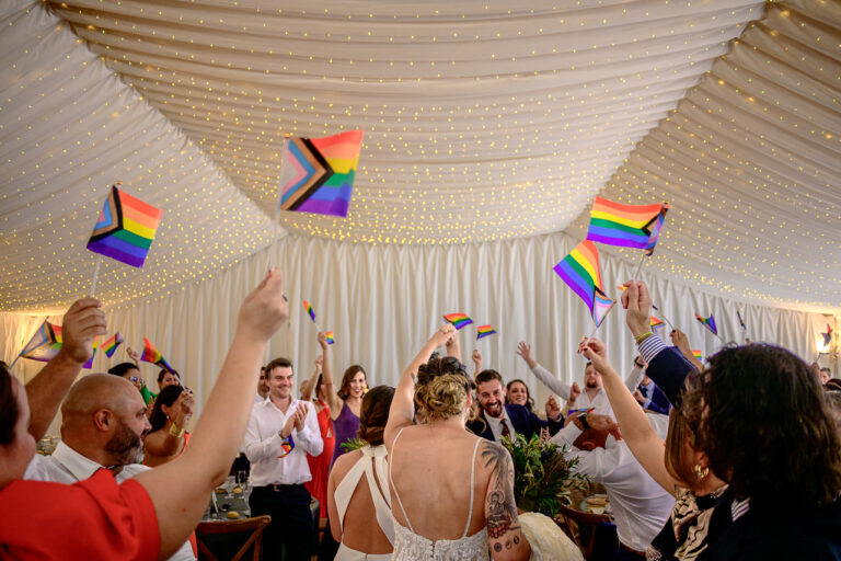 Fotógrafo de bodas natural y sin posados | Adolf Boluda 10 Dos novias entran al salón recibidas por todos los invitados ondeando banderas LGBTQ+, boda natural en Finca el Vincle, Alicante.