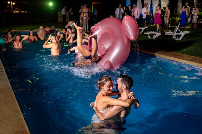 Fotógrafo de bodas natural y sin posados | Adolf Boluda 8 Los novios se abrazan en la piscina mientras, al fondo, sus amigos vuelcan un flotador flamenco entre risas en plena fiesta nocturna en Énova, Valencia.
