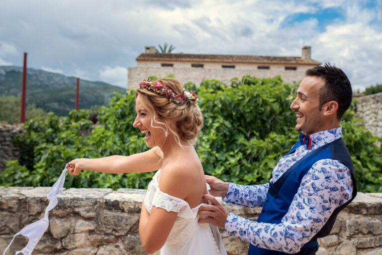 Fotógrafo de bodas natural y sin posados | Adolf Boluda 7 El novio abrocha el vestido a la novia desde atrás mientras ella gira riendo el sujetador que acaba de quitarse, preparativos en Casa Benigalip, Alicante.