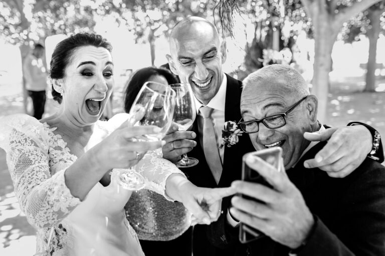 Fotografía de bodas documental: qué es | Adolf Boluda 7 Novia y familia riendo durante una videollamada con un familiar que no pudo asistir, en el cóctel, boda documental en La Masia de Chencho